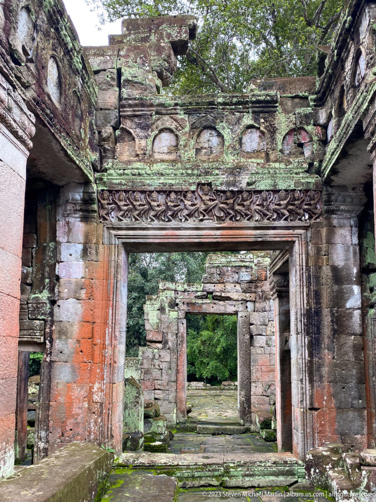 Roofless sandstone doorway beneath a carved figure frieze and blind arches, with ruins beyond.