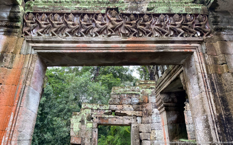 Row of small carved figures above a sandstone doorway opening toward ruins and forest.