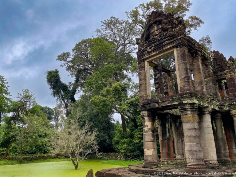 Tower-like end of a columned sandstone building beside a green pond and forest.