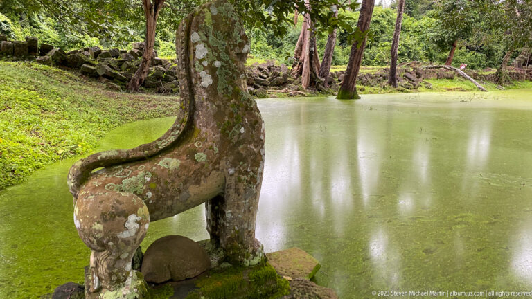 Rear view of a weathered sandstone lion guardian beside a green temple pond.