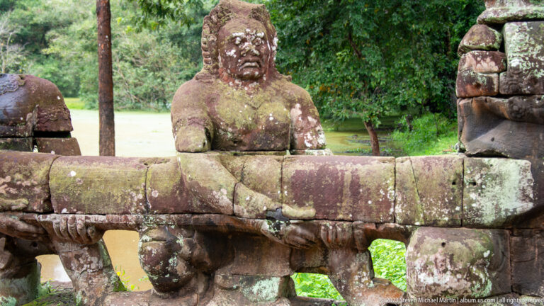 Weathered stone guardian with hands beneath a horizontal serpent-form balustrade.