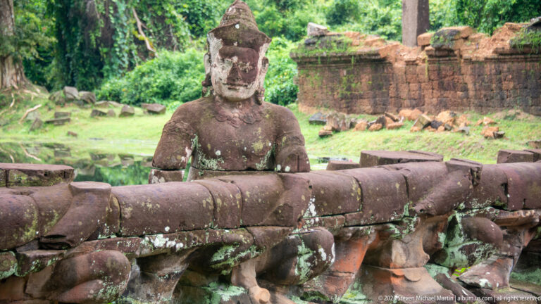 Frontal stone guardian with headdress behind a thick carved serpent-form railing.