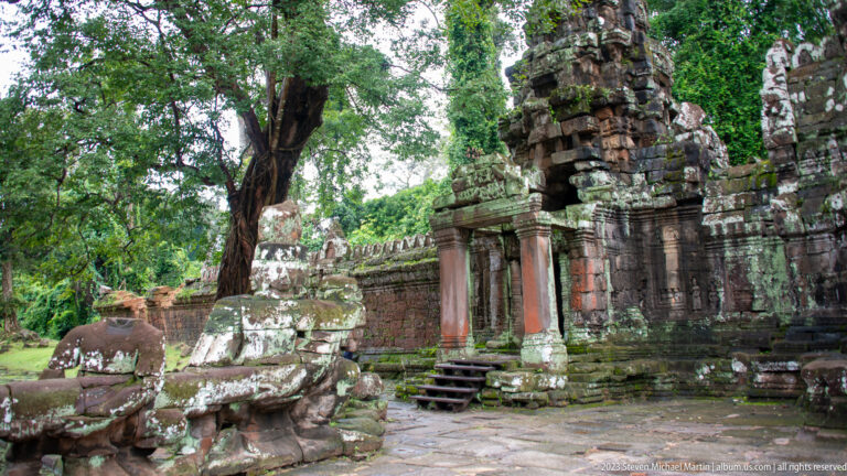 Side court with a small temple doorway, columns, stone steps, fragments, and a large tree.