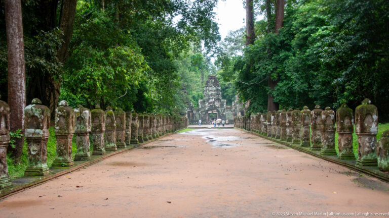 Straight temple causeway lined with stone guardian figures and ending at a distant tower among trees.