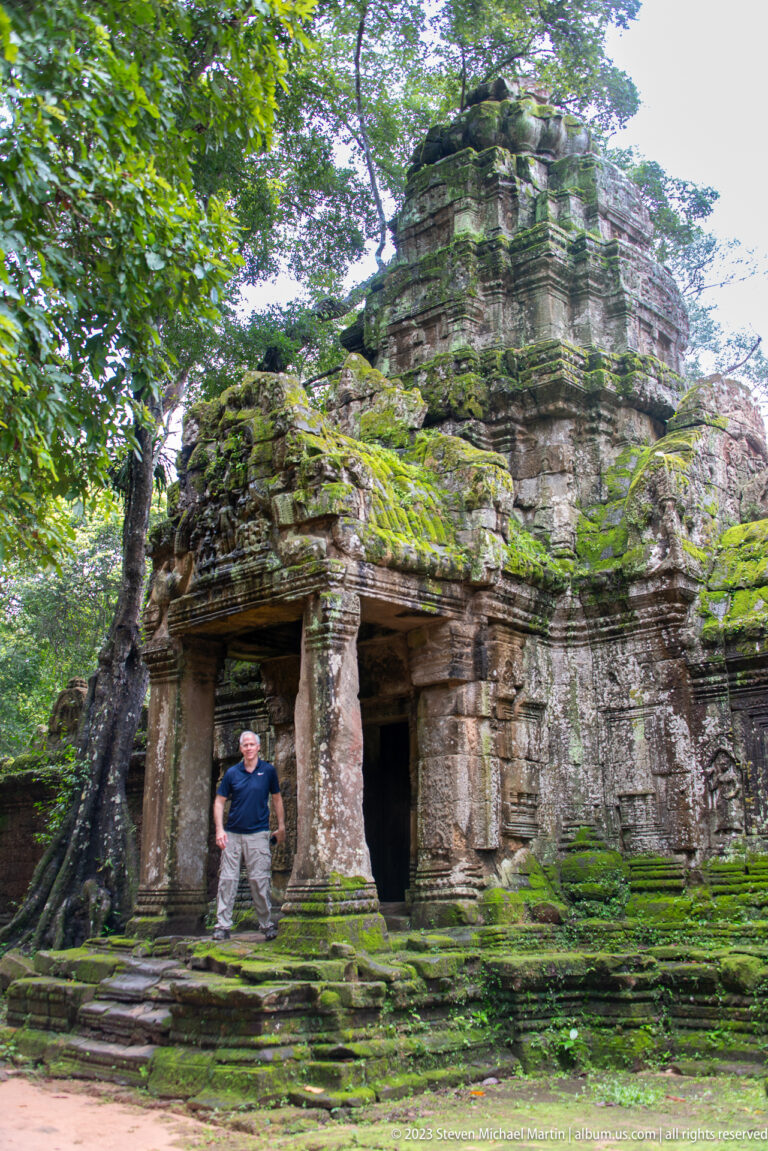 Visitor standing before a mossy temple doorway beneath heavy tree roots and weathered stonework.