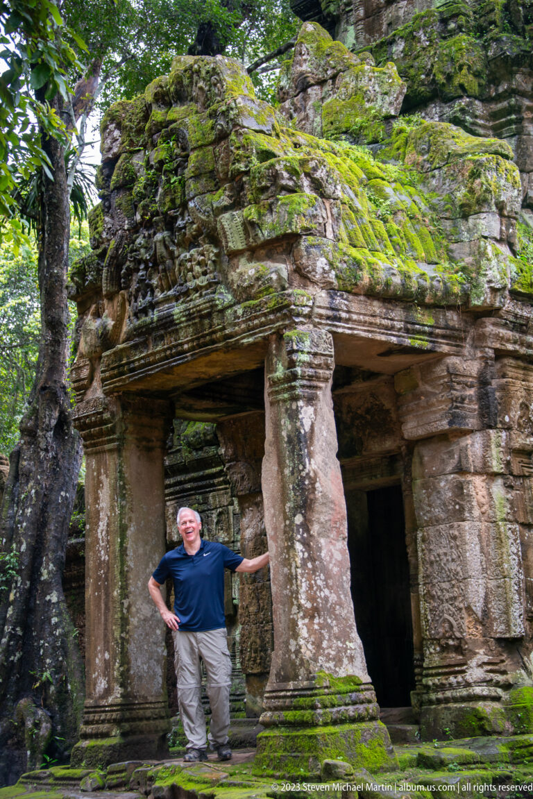 Visitor beside a worn temple column under thick roots and a moss-covered stone façade.