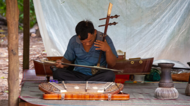 Seated musician playing a bowed string instrument beside a trapezoidal hammered dulcimer.