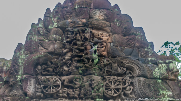 Arched Khmer pediment densely carved with a central deity, attendants, and curling vegetal ornament.