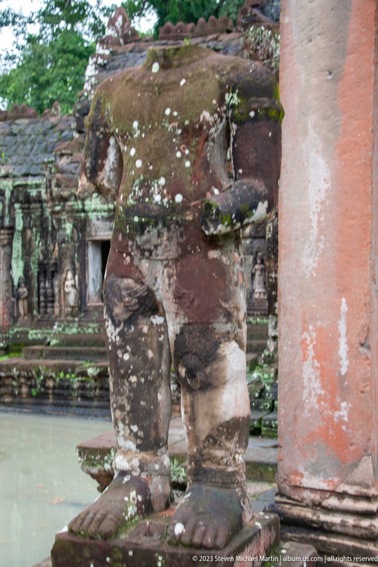 Lower half of a damaged standing stone guardian beside a doorway column on a round base.
