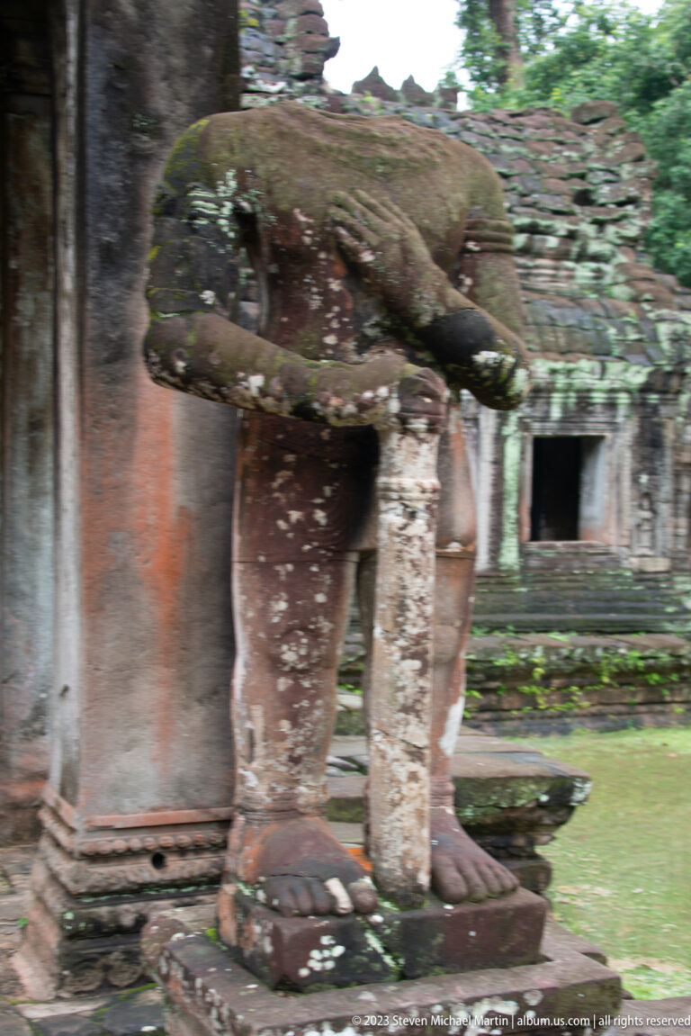 Side view of a broken stone guardian with bent body and large feet beside a temple entrance.