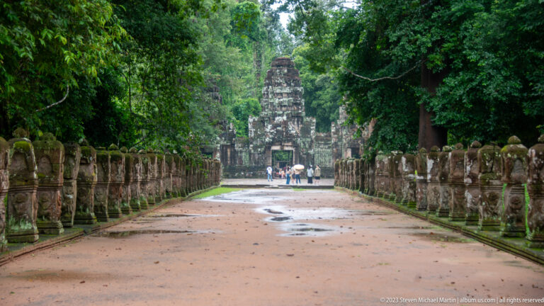 Closer view down a reddish causeway lined by stone figures toward a tall temple entrance.