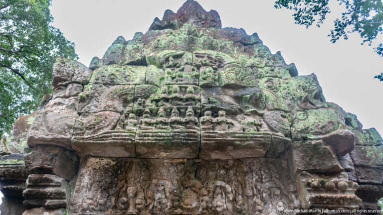 Low-angle view of a large arched Khmer pediment covered with figures and curling stone ornament.