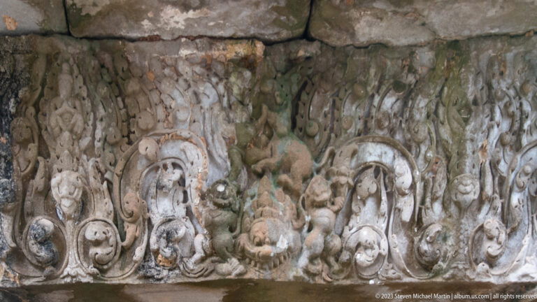 Close view of a weathered stone frieze with curling leaves, floral motifs, and small carved figures.