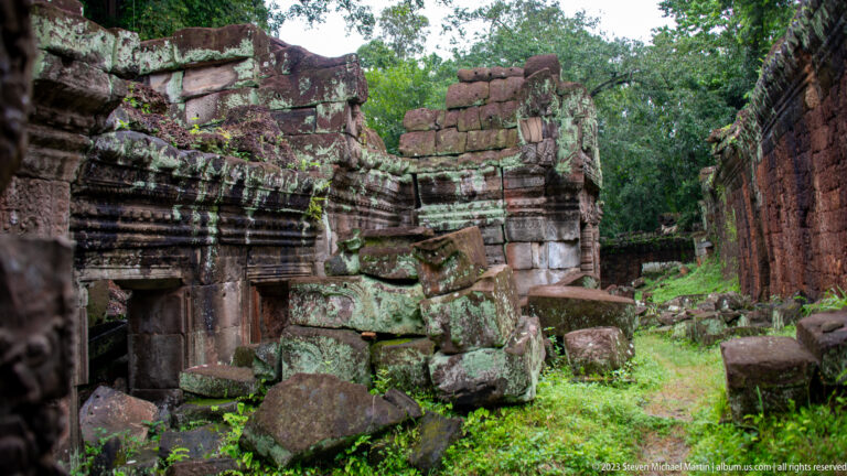 Ruined temple passage with fallen stone blocks, standing carved walls, and grass growing between them.