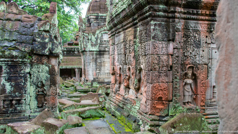 Oblique view of a carved temple wall with niches, relief panels, and fallen stone blocks beside it.