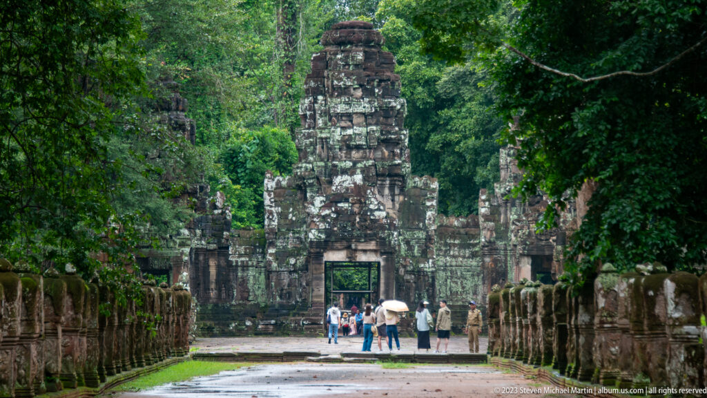 Tall stone temple entrance centered beyond rows of guardian sculptures, with visitors at the doorway.