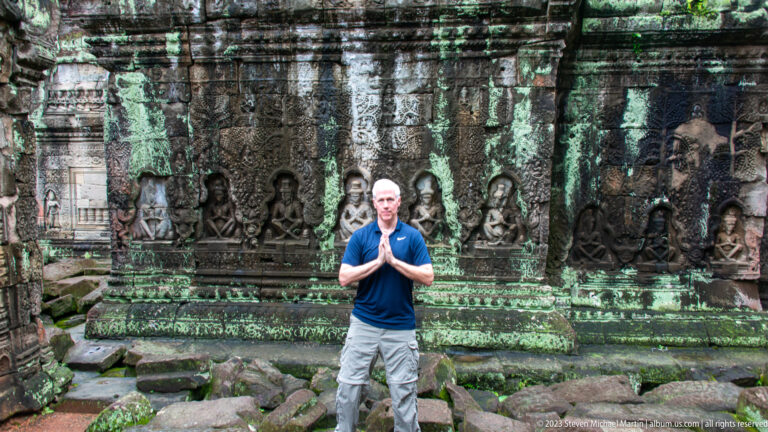 Visitor with palms together standing before a wall of seated figures in carved niches.