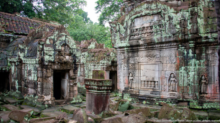 Ruined temple court with adjoining shrines, a decorated wall panel, central stone element, and fallen blocks.