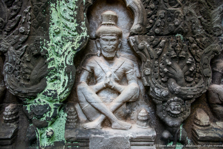 Seated stone figure with crossed legs inside an ornate arched niche of curling foliage.