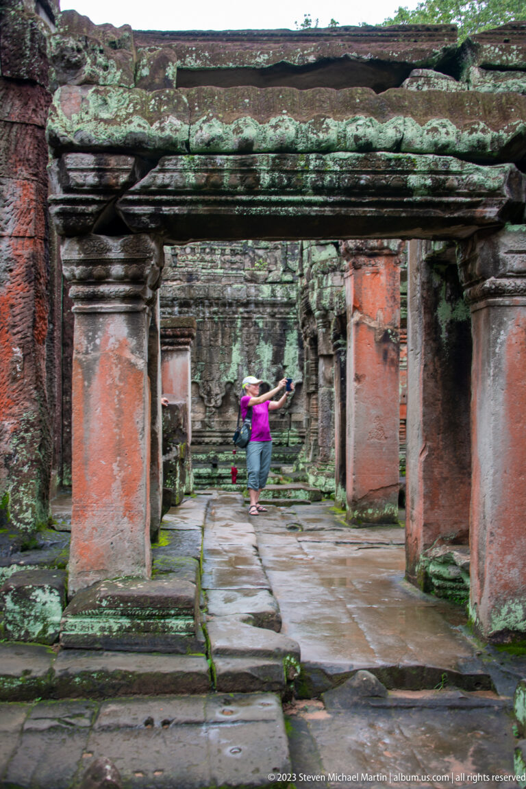 Visitor walking through a long temple corridor of stone columns, lintels, and aligned openings.