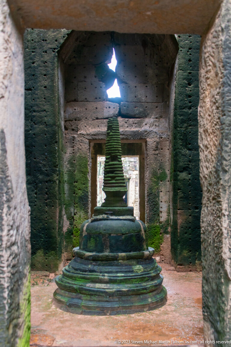Centered tiered stone monument on a round base inside a backlit chamber with green surface growth.