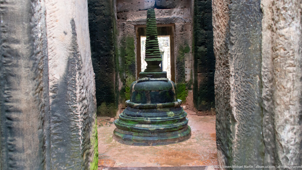 Small tiered stone stupa centered in a narrow sandstone corridor with aligned doorways behind it.
