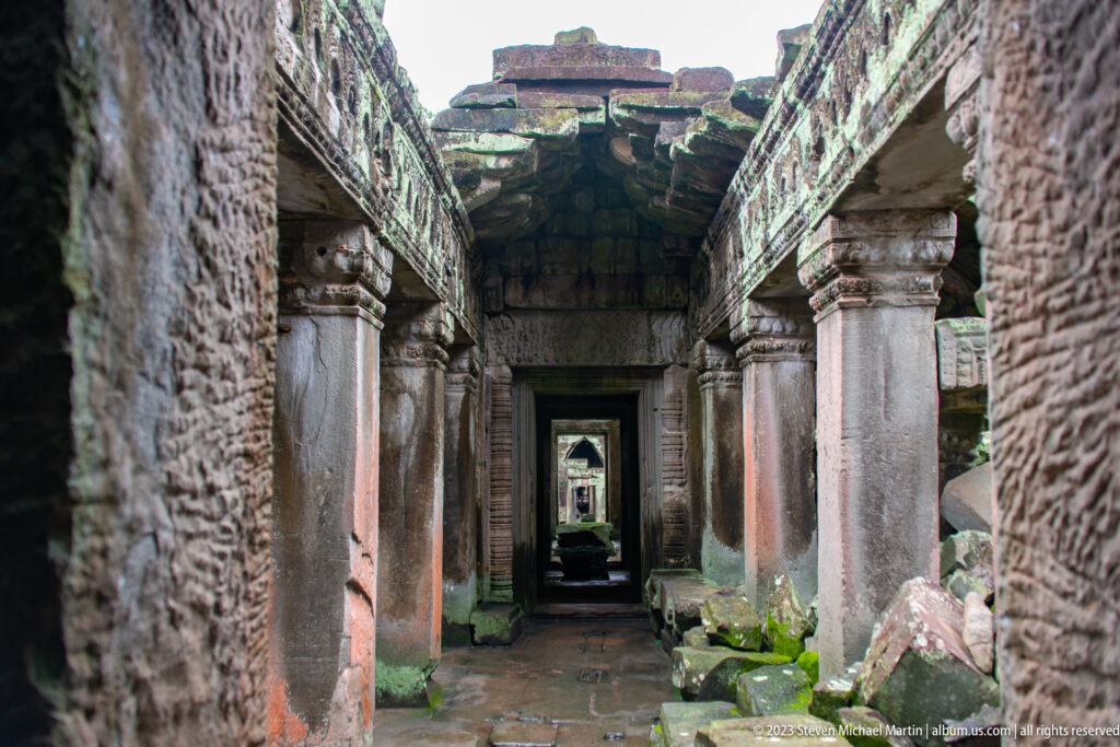 Weathered sandstone corridor with square columns, scattered blocks, and several aligned doorways.