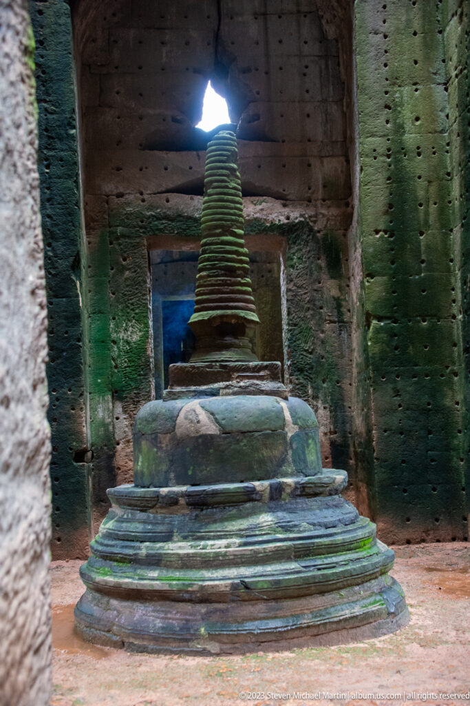 Tiered stone stupa in a dark chamber beneath a bright opening in the collapsed roof.