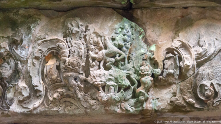 Weathered sandstone lintel with a central monster face and curling vegetal relief on both sides.