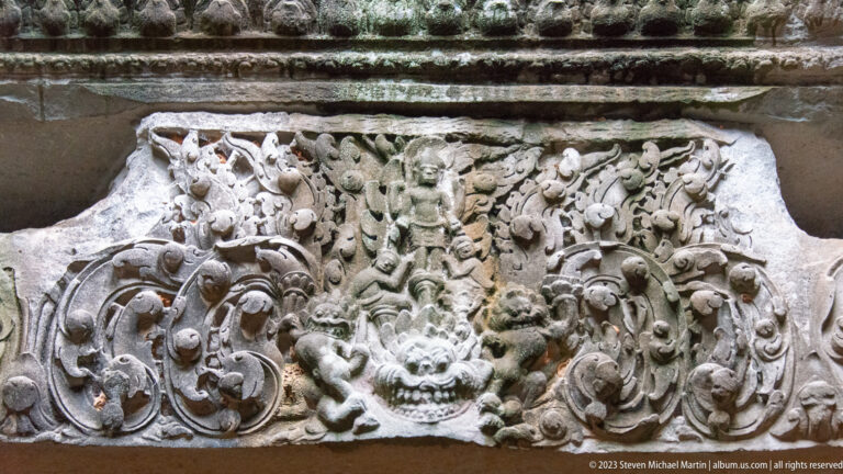 Rectangular sandstone lintel carved with a central face, small figures, and symmetrical curling foliage.