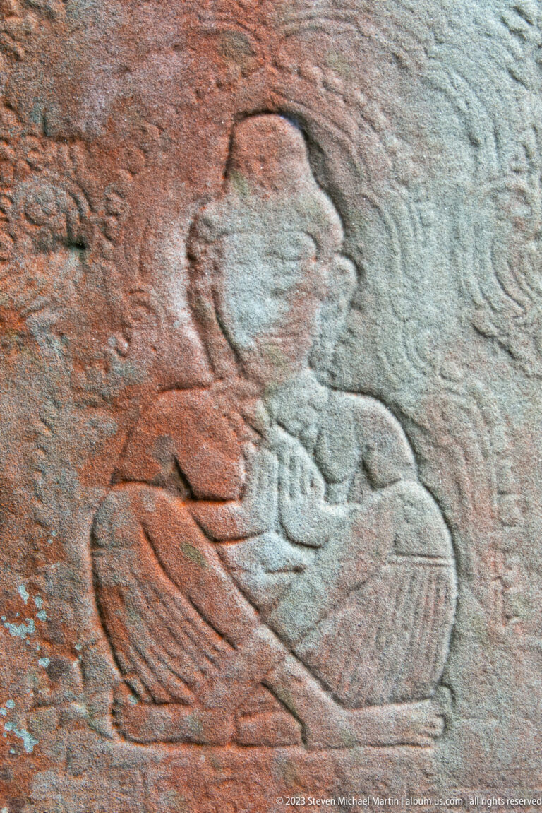 Faint sandstone bas-relief of a seated figure holding an object against the torso.