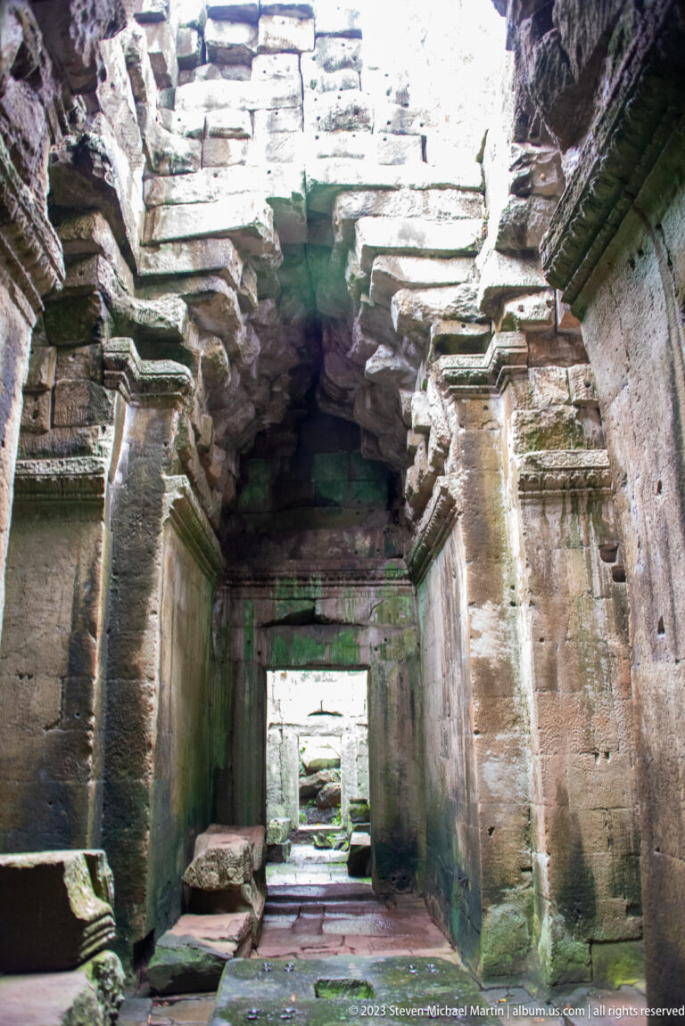Collapsed sandstone chamber with carved pilasters, fallen roof blocks, and a doorway centered beyond.