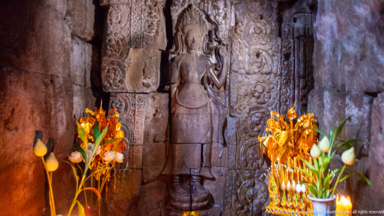 Carved standing female figure beside an ornate doorway with bright floral offerings in the foreground.