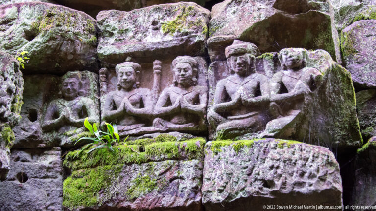 Five kneeling sandstone figures with hands joined in prayer, surrounded by moss and broken carving.