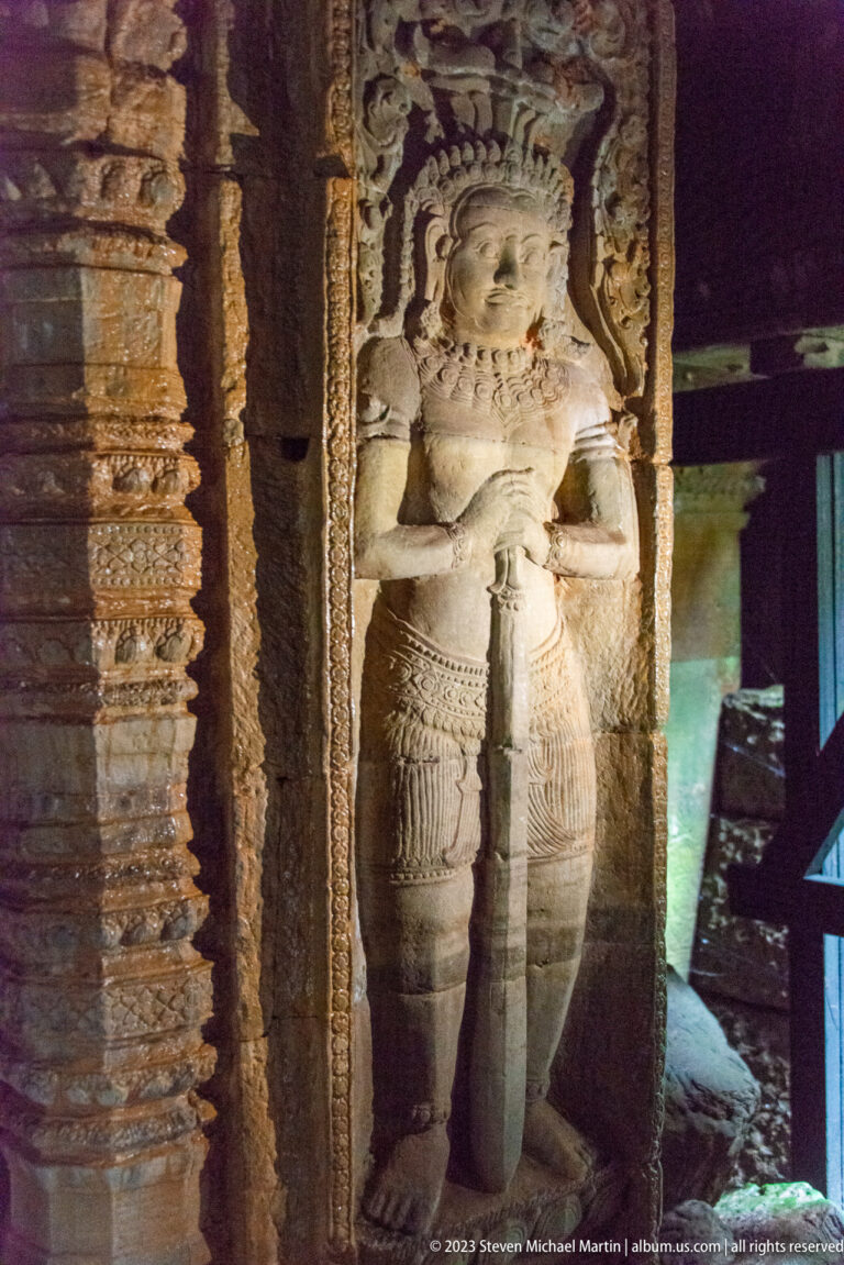 Large standing female deity with elaborate crown and jewelry carved beside an ornate doorway.