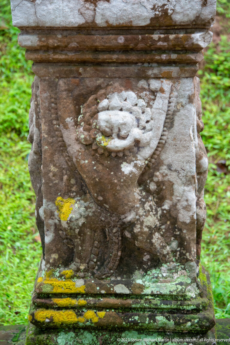 Weathered guardian relief on a sandstone pedestal with green and yellow moss staining.