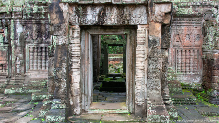 Ornate sandstone doorway flanked by carved figures and baluster windows, opening into a ruined chamber.