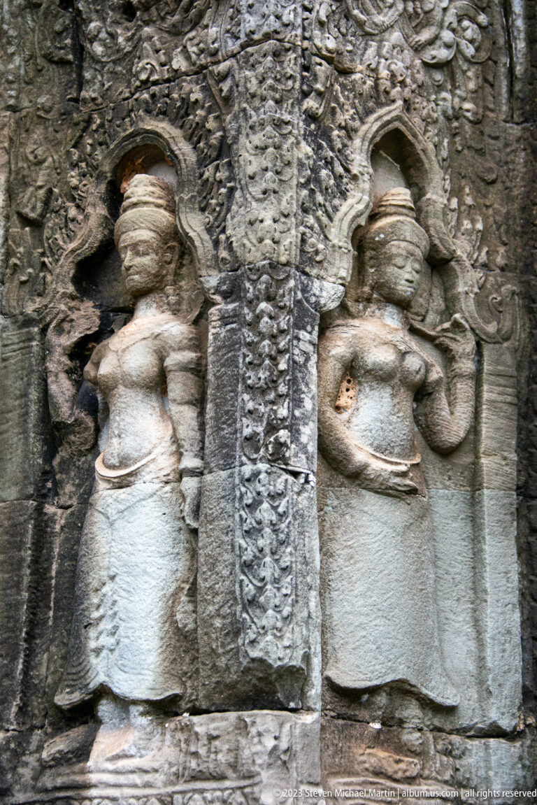 Two symmetrical standing female deities carved on sandstone beside a narrow vegetal border.