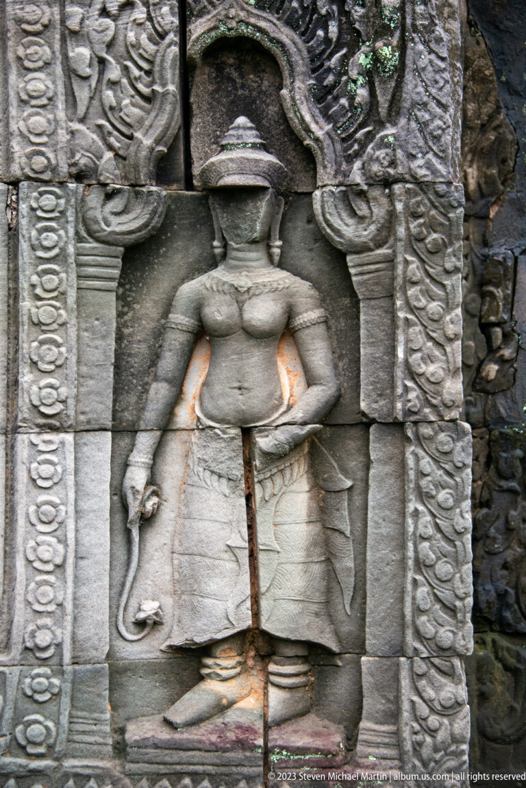 Standing female deity in a carved niche holding a long-stemmed object, with a vertical crack through the stone.