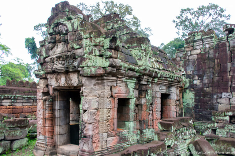 Small lichen-covered sandstone shrine or gateway with several openings and a partly collapsed roofline.