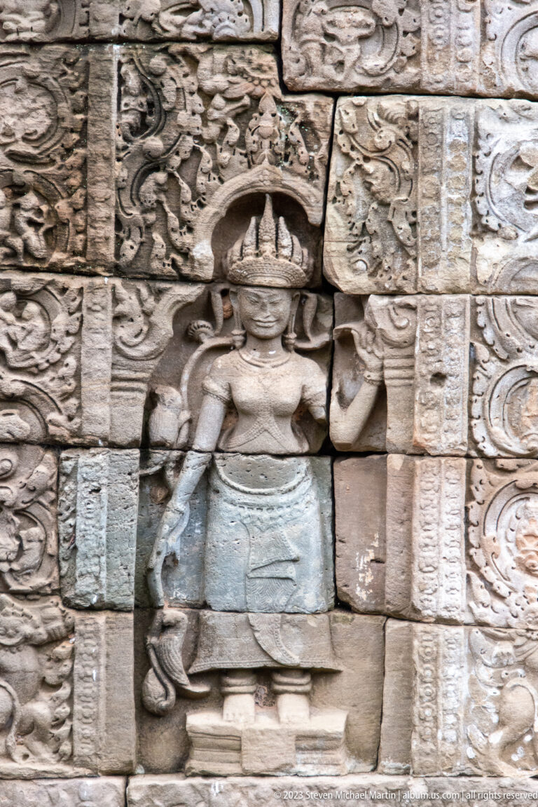 Standing female deity with flared headdress and raised hand carved amid dense sandstone ornament.
