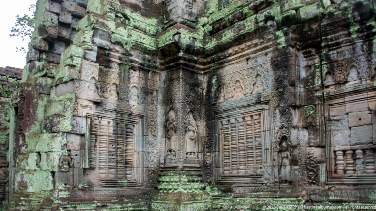Lichen-covered sandstone interior wall with baluster windows, carved panels, figures, and corner moldings.