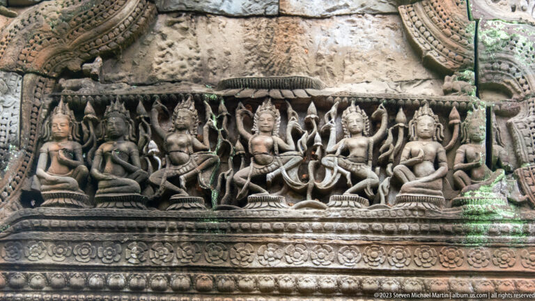 Sandstone frieze of seated crowned figures beneath a scalloped border and above floral ornament.