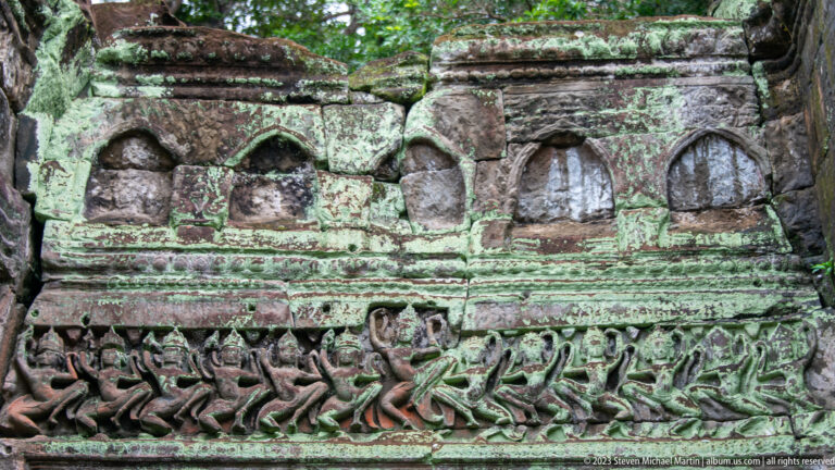 Moss-covered sandstone frieze with repeated small figures beneath a row of blind arched niches.