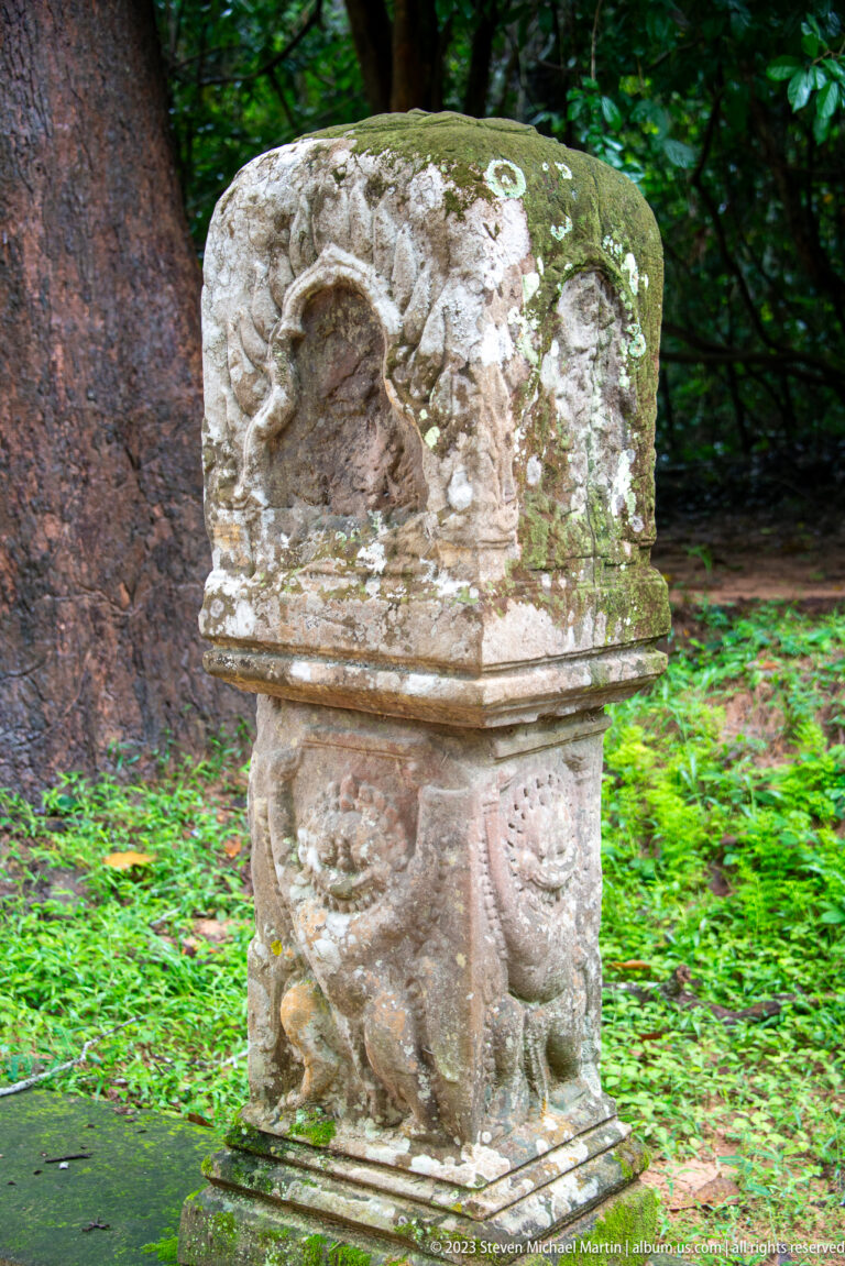 Damaged carved sandstone pedestal with lichen, a broken upper figure, and paired lower motifs.
