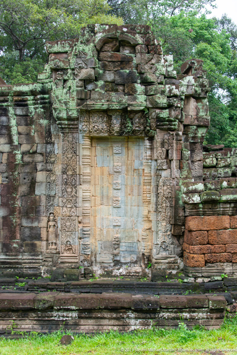 Tall sealed sandstone doorway framed by carved pilasters in a partially collapsed temple façade.