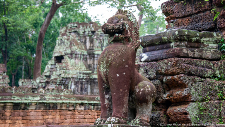 Profile of a weathered sandstone lion guardian with lichen and ruined temple walls behind.