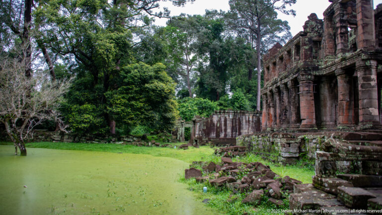 Two-story sandstone building beside a green duckweed-covered pond with fallen blocks at its base.