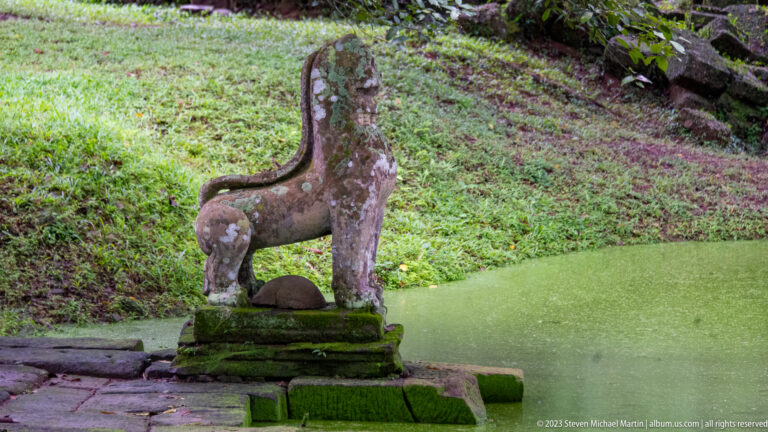 Weathered sandstone lion guardian on a pedestal beside a green temple pond.