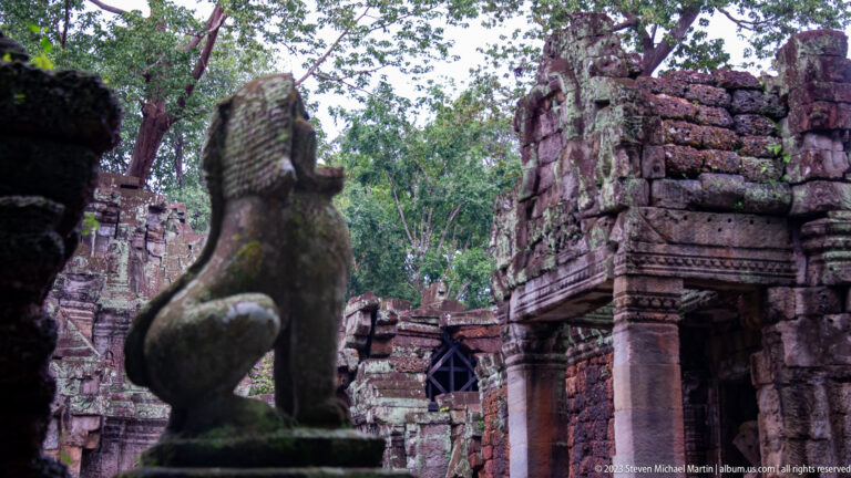 Profile of a sandstone lion guardian in the foreground with ruined temple towers behind.